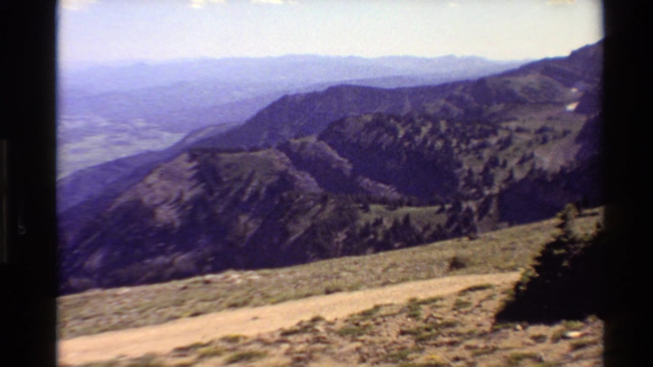 1980 Western USA home-movie frame with a pale dirt path, dark evergreen trees, rounded ridges, and hazy valley layers in the distance.