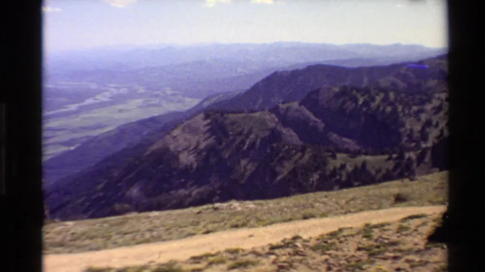 1980 Western USA 8mm frame looking across a dirt trail and rugged mountain slope toward a broad, hazy valley floor.