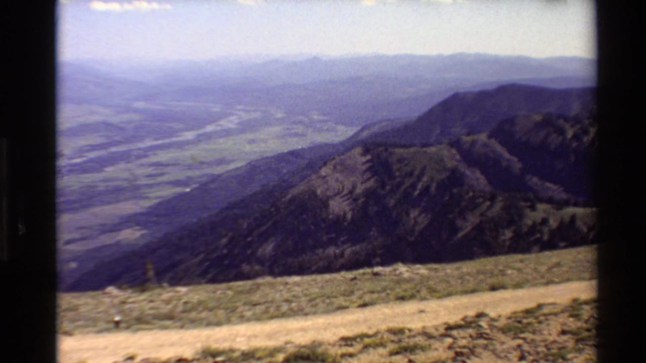 1980 Western U.S. 8mm frame showing a hazy mountain valley from an elevated overlook, with ridges, a river, scrubby ground, and a dirt path.