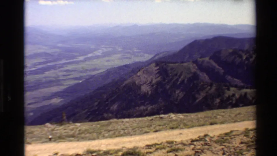 1980 Western U.S. 8mm frame showing a hazy mountain valley from an elevated overlook, with ridges, a river, scrubby ground, and a dirt path.