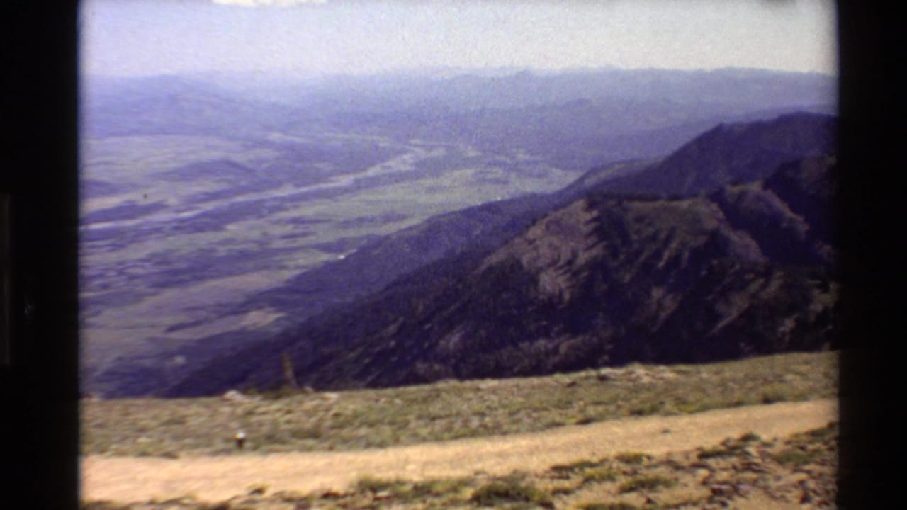 Panoramic mountain valley view with a river, dark ridge, pale foreground path, and hazy distant hills.