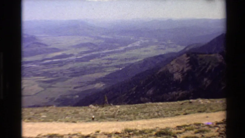 1980 Western U.S. 8mm still of a high overlook toward a hazy valley floor, with layered slopes and a river visible in the distance.