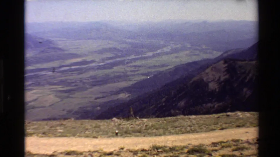 1980 Western U.S. landscape frame from a mountain overlook, showing a distant river valley, sloped ridge, scrub foreground, and hazy sky.