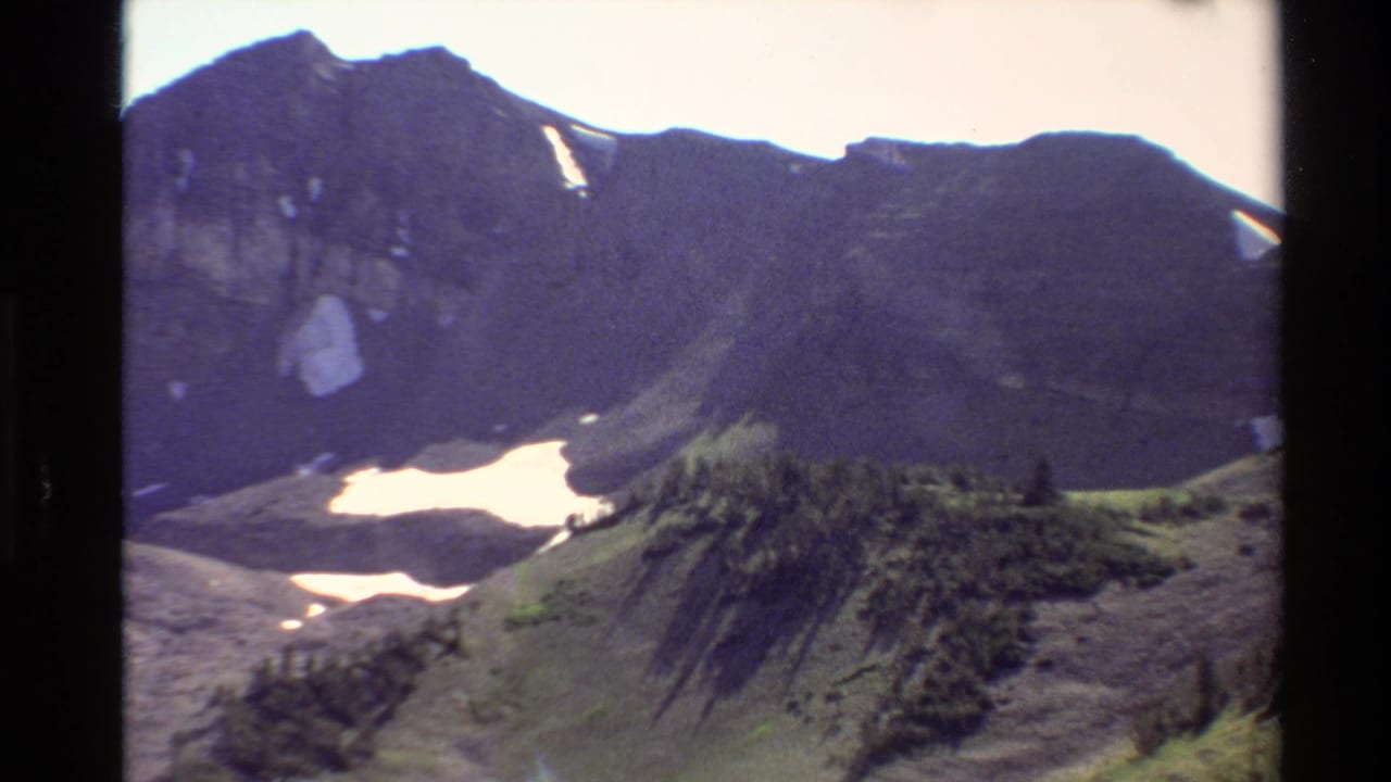 Western USA 1980 8mm frame showing a dark alpine ridge, small snowfields, green slopes, and evergreen trees.