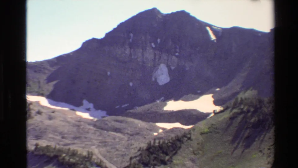 Western USA 1980 8mm frame showing a rocky mountain face, remaining snow, green alpine terrain, and scattered trees.
