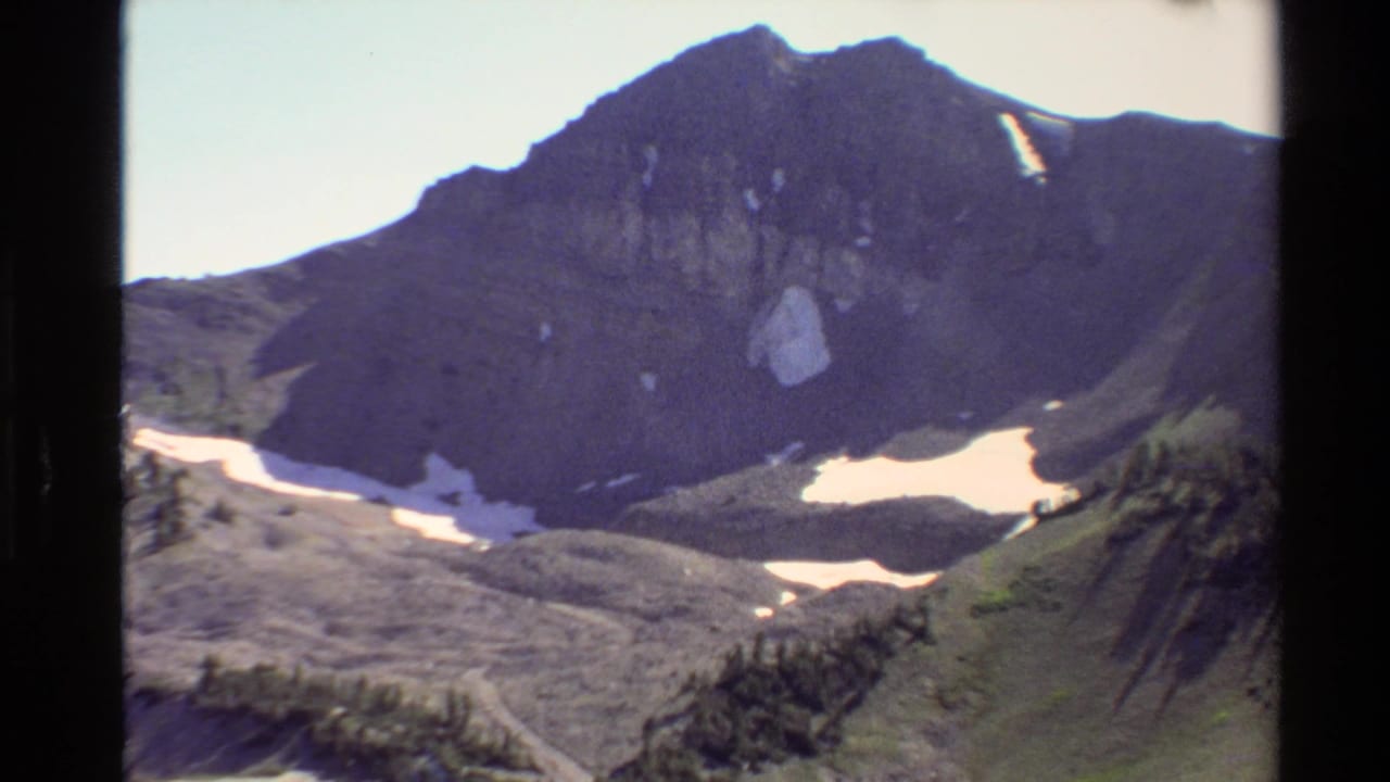1980 Western U.S. 8mm frame of a dark rocky mountain ridge above snow patches, sparse trees, and open alpine terrain.