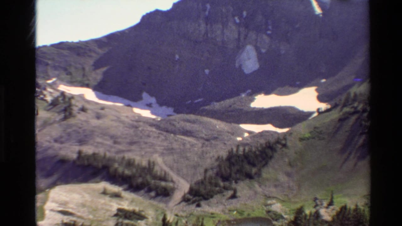 Mountain basin with snow patches, rocky slopes, trees, and a winding path.