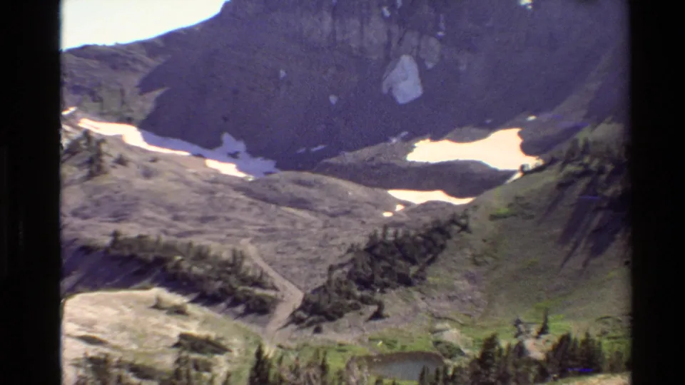 1980 Western U.S. 8mm frame looking toward rocky alpine slopes, residual snow, tree clusters, and a small dark water area near the lower edge.