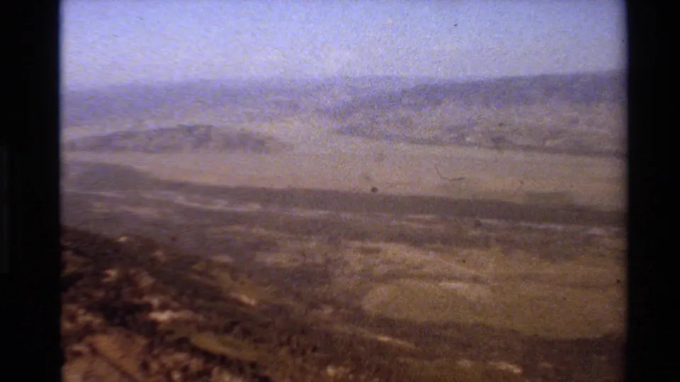 1980 Western USA 8mm frame showing a hazy mountain valley, dry foreground slope, and pale sky during a handheld panorama.