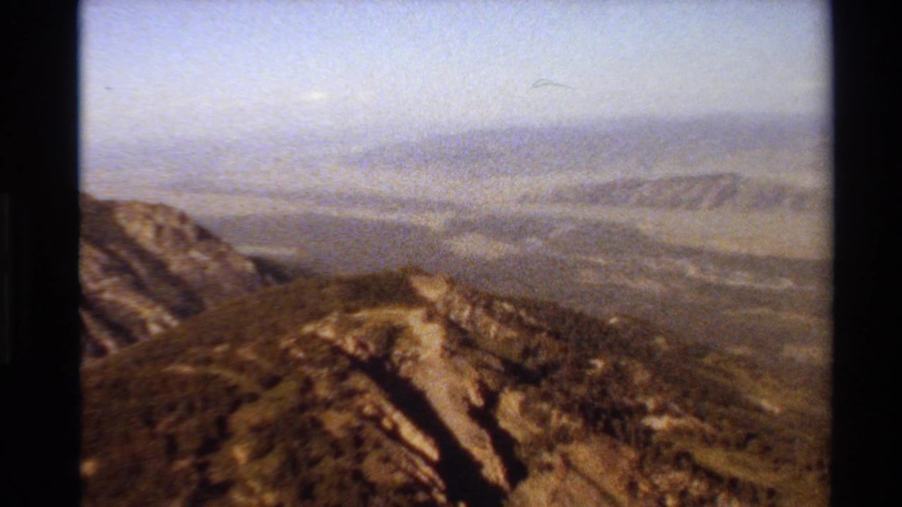 1980 8mm Western U.S. mountain view with a rocky foreground ridge, tree-covered slopes, and hazy distant terrain under pale sky.
