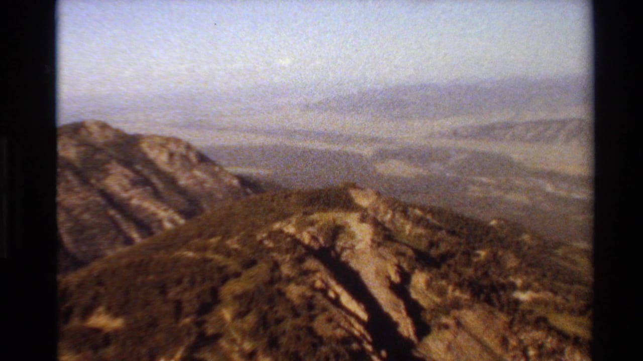 Western U.S. 8mm landscape frame showing a rugged ridge, sloped rocky ground, distant mesas or mountains, and a hazy sky.