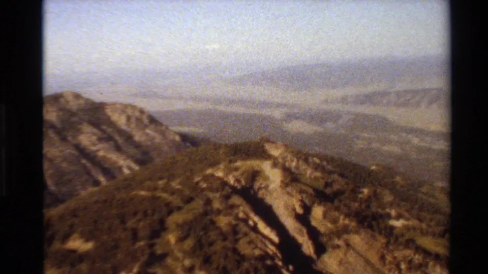1980 home-movie frame of a Western U.S. mountain overlook with rocky ridges, sparse tree cover, distant valley forms, and faded color.