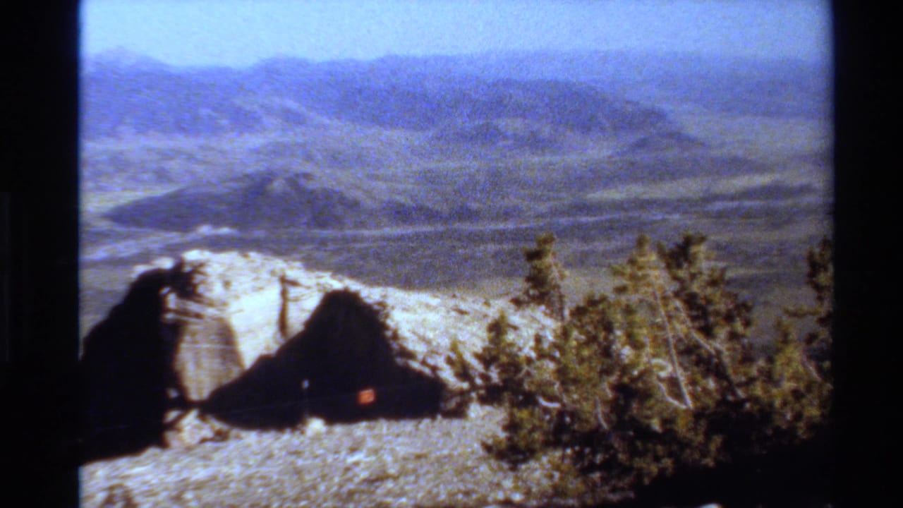 1980 Western USA 8mm frame showing pale rocks, sparse brush, a broad valley, and mountain ridges under blue sky.