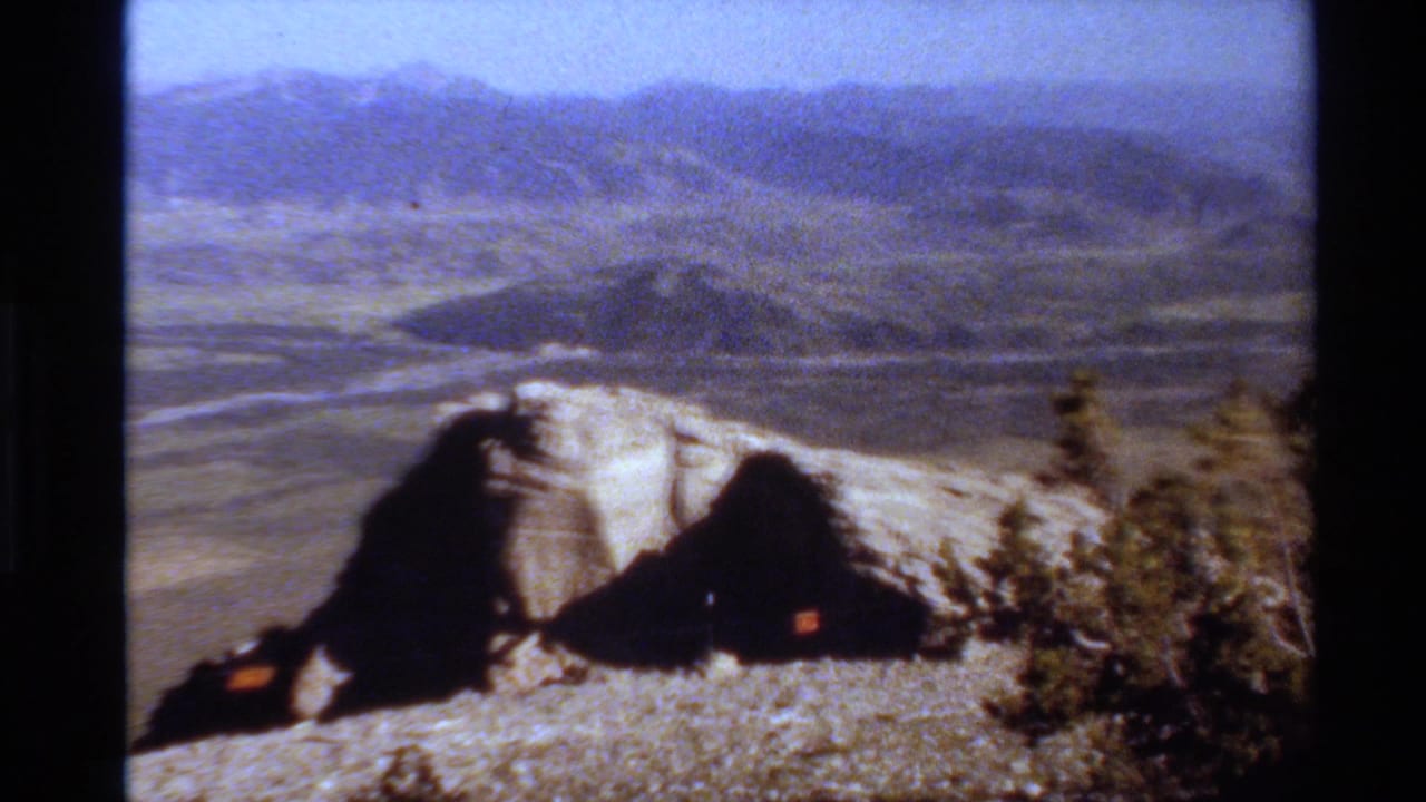 Western USA home-movie frame with rocky foreground, small dark shelter-like shapes, open valley, and distant ridges.