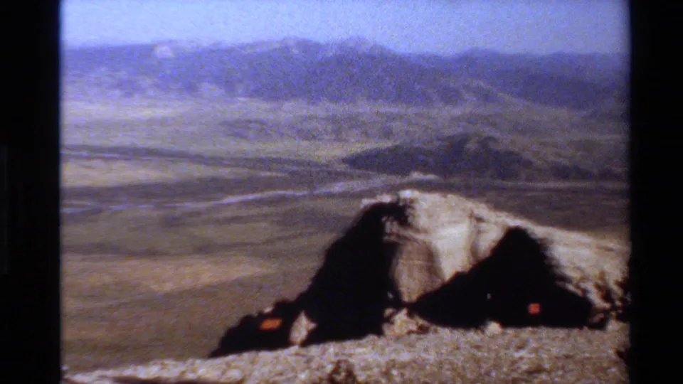 1980s 8mm landscape frame from a high overlook, with rocks in front and hazy mountain terrain across the valley.