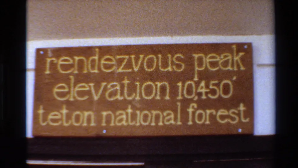 8mm home-movie frame showing the Rendezvous Peak elevation 10450 sign mounted between pale posts in Teton National Forest.