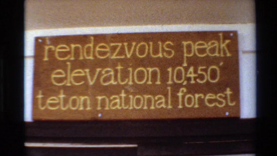 1980 home-movie frame of the wooden Rendezvous Peak elevation sign in Teton National Forest, softly focused with visible film grain.
