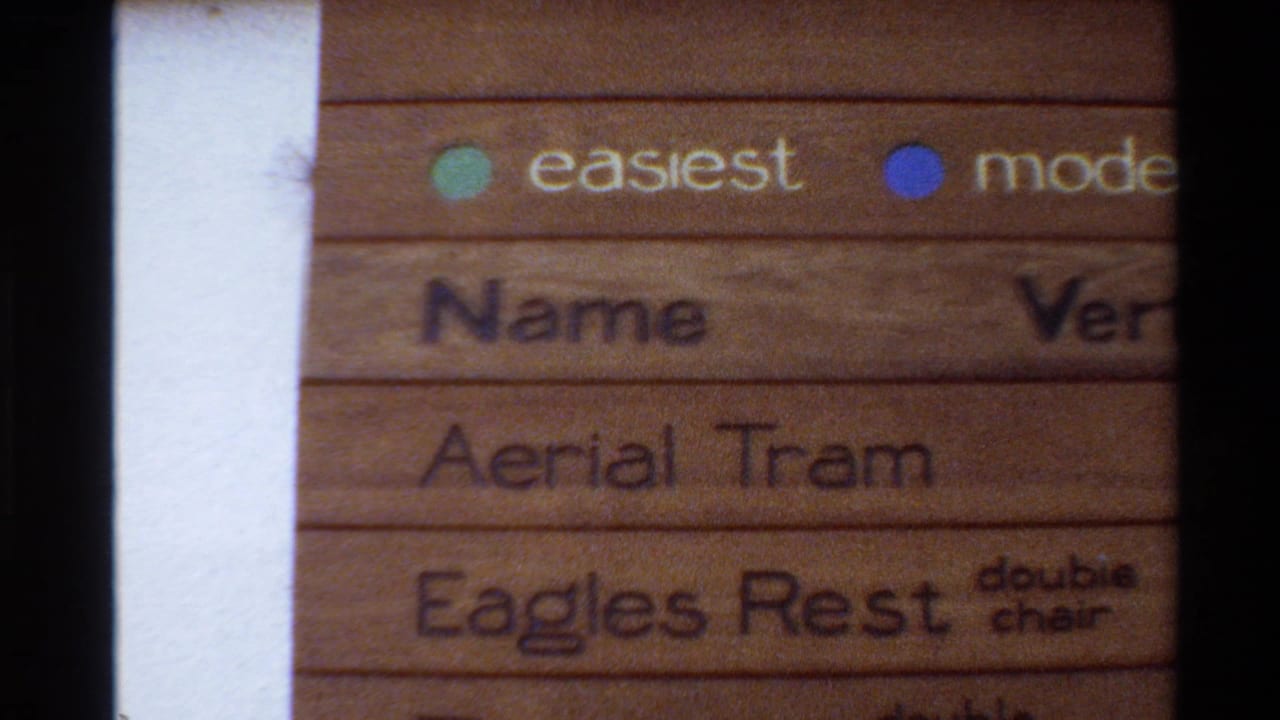 1980 Western USA 8mm frame showing a close handheld view of a wooden mountain resort sign with trail difficulty dots and partial lift listings.
