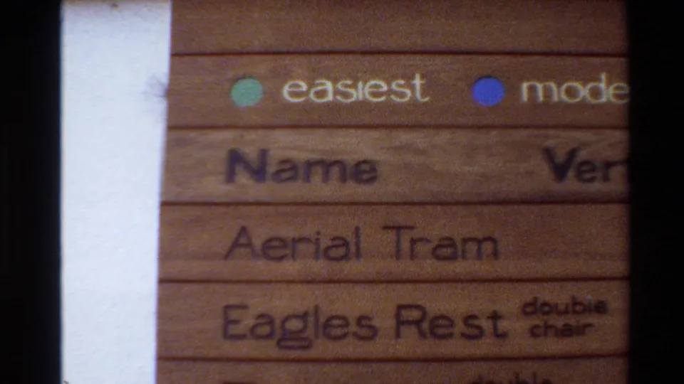 1980 Western USA 8mm frame showing a close handheld view of a wooden mountain resort sign with trail difficulty dots and partial lift listings.