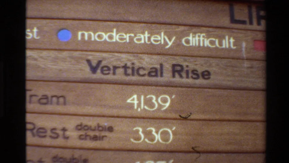 Handheld 8mm frame of the resort wayfinding sign, with difficulty labels and vertical-rise numbers for tram and chairlift entries.