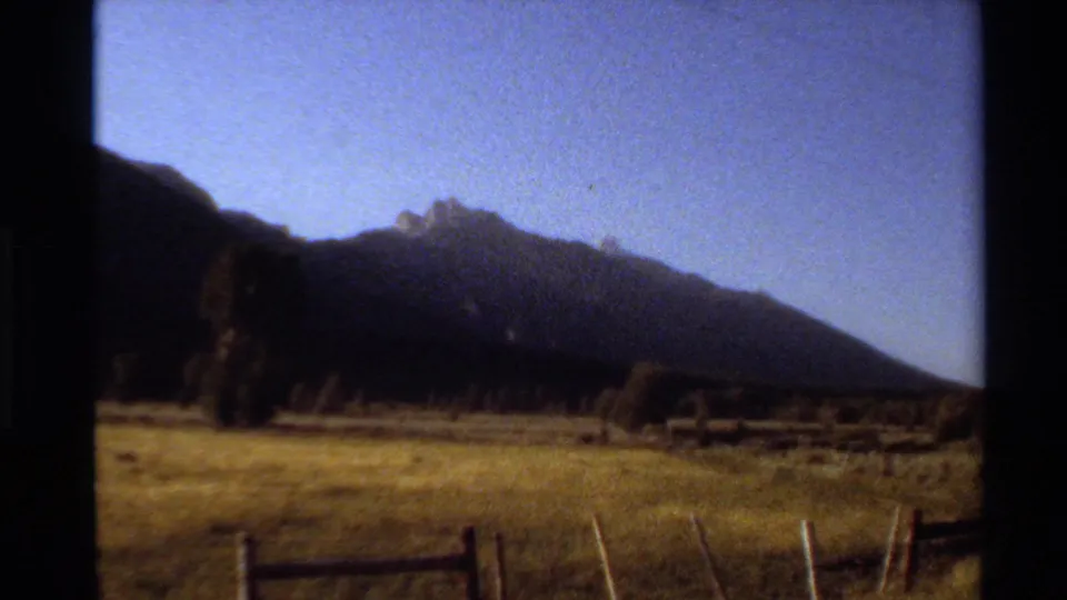1980 Western U.S. 8mm frame showing a mountain ridge beyond a grassy field, scattered trees, clear sky, and foreground fencing.