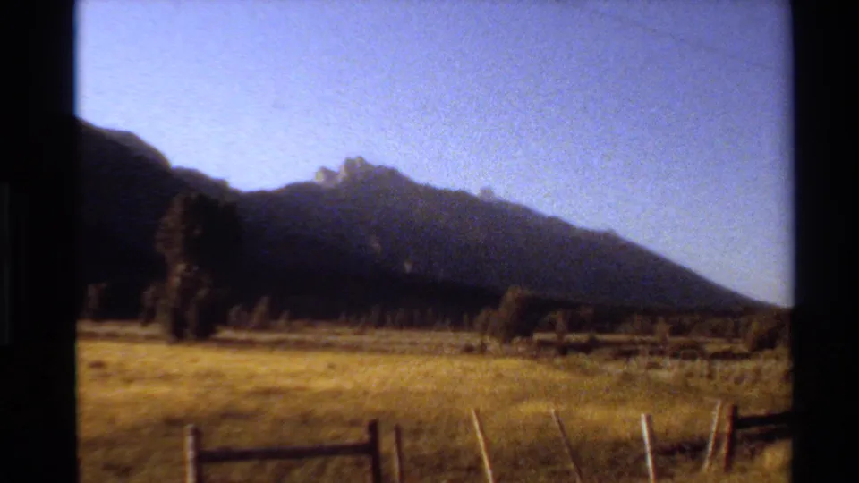Western U.S. 1980 home-movie frame of a mountain landscape with open grass, trees, and a fence in the foreground.