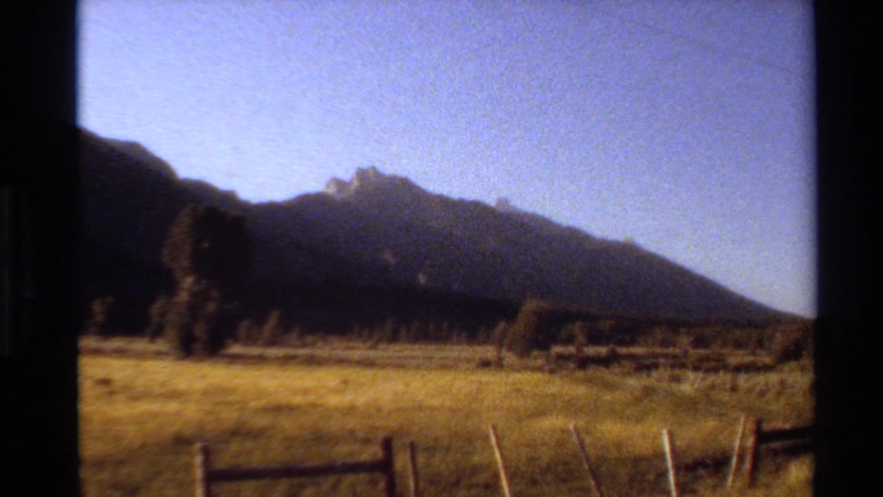 1980 Western USA 8mm frame showing grassland, fence rails, and a dark hill or mountain ridge beneath clear sky.