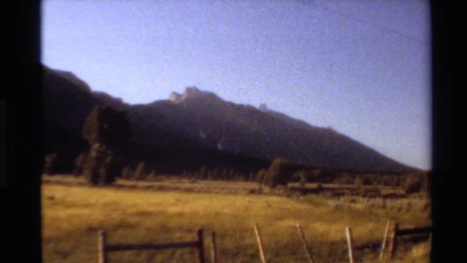 Western USA home-movie frame from 1980 with a fenced grassy field leading toward a dark hill or mountain ridge.