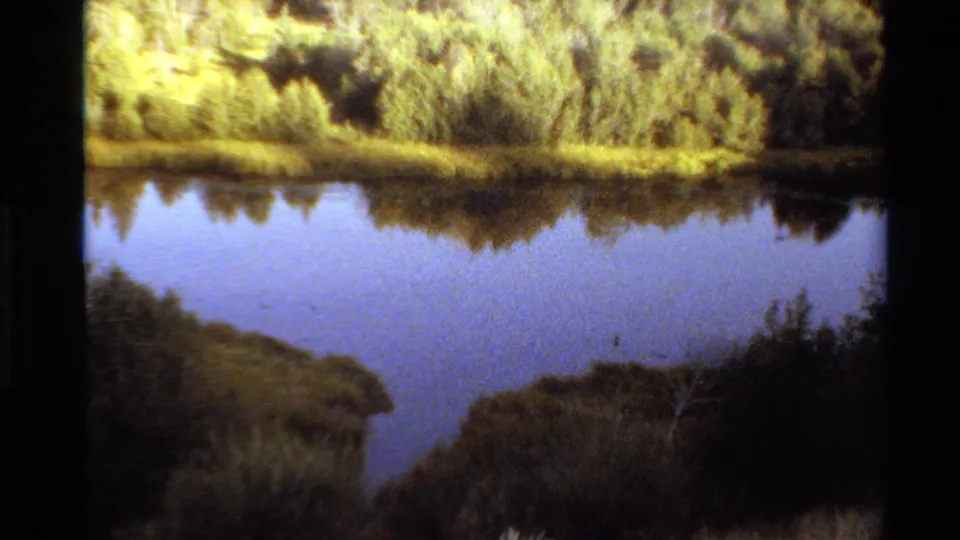 1980 Western USA 8mm frame showing still water bordered by shrubs and grass, reflecting the sky and tree line.