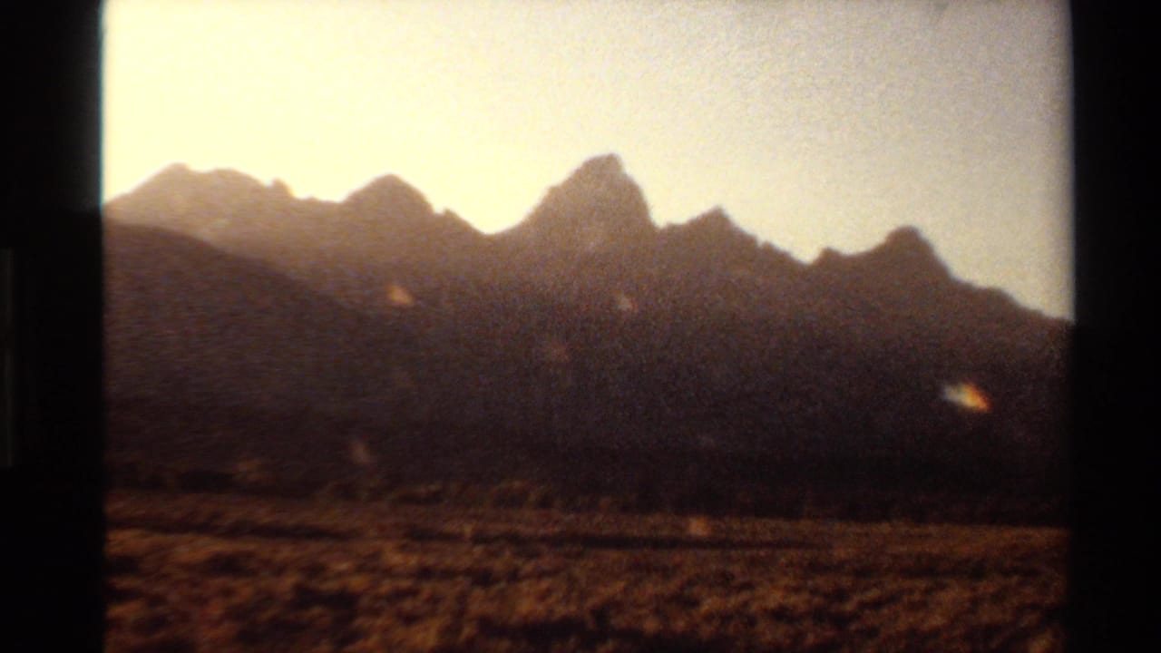 1980 Western USA 8mm frame showing jagged mountains silhouetted beyond dry grassland in warm late-afternoon light.