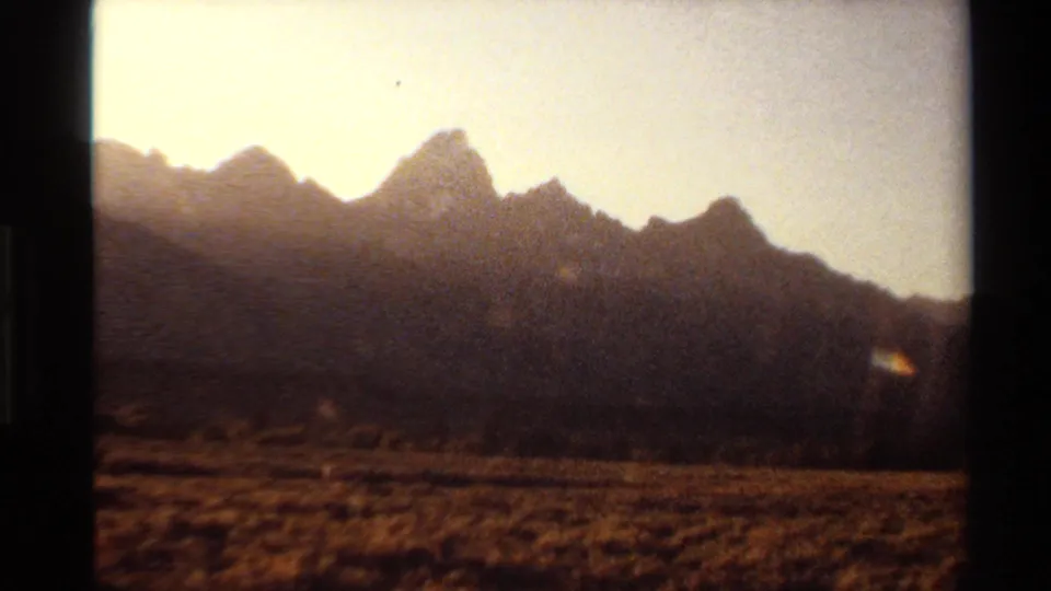 1980 Western USA home-movie frame with a rugged mountain silhouette, dry grassland, warm sky, and small lens flare near the ridge.