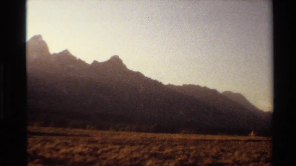 1980 Western USA home-movie frame with a long mountain slope, dry grassland foreground, and bright late-afternoon sky.