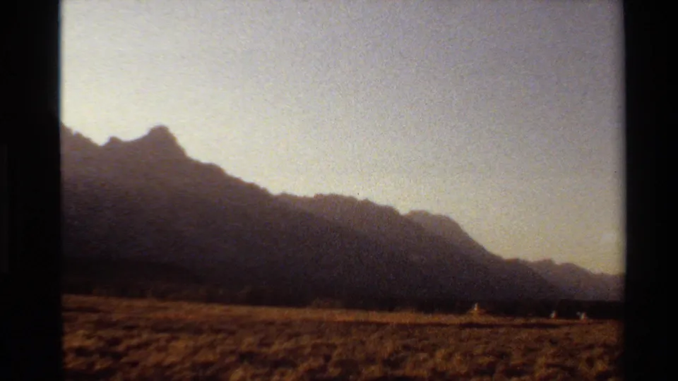 Western U.S. 8mm frame showing silhouetted mountains, dry grassland, and small distant people in sunset light.