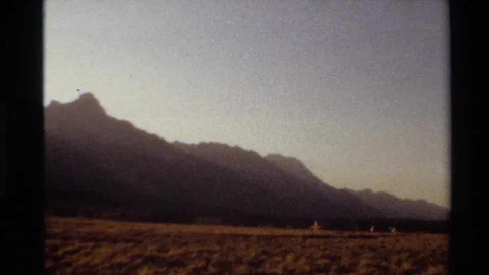 Home-movie frame from the Western U.S. with a dark mountain slope, open grassland, and distant people at sunset.