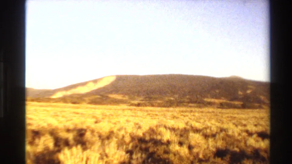 Warm-toned 8mm frame of dry grassland or scrub leading to low mountains under a bright sky in Western U.S. travel footage.