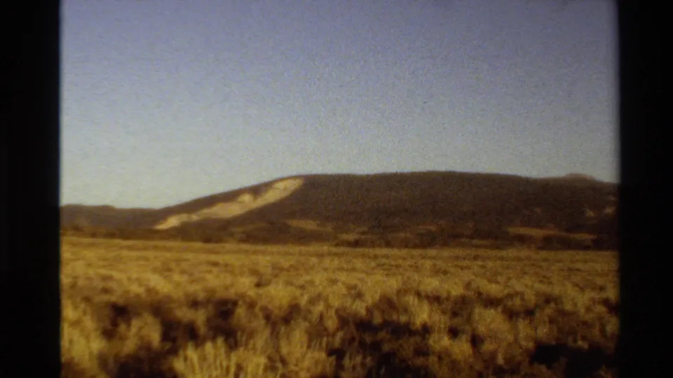 1980 8mm landscape frame showing scrubby open ground, a distant ridge, and muted sky with soft grain and warm exposure.