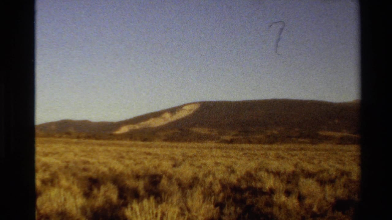 1980 Western U.S. 8mm frame of dry sagebrush terrain leading to a distant mountain ridge beneath a clear sky.