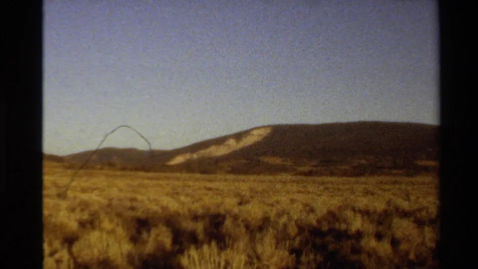 Vintage 8mm view of a Western U.S. high desert field, with golden scrub in front of a low mountain ridge.