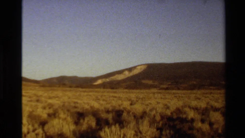1980 home-movie frame showing sagebrush or dry scrub across open land below a distant mountain slope.