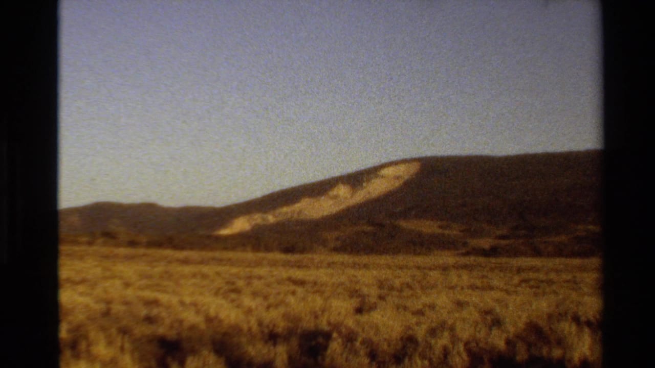 1980 Western U.S. 8mm frame showing a dry open field, dark ridge, and light-colored exposed slope beneath a pale sky.