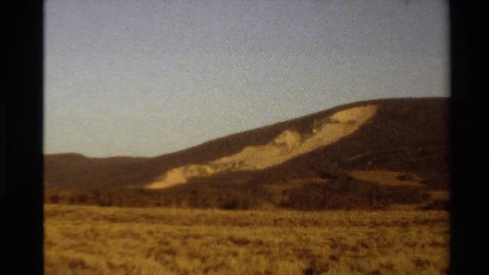 1980 Western U.S. 8mm frame showing a closer hillside view, dry foreground grass, and a pale exposed slope across the ridge.