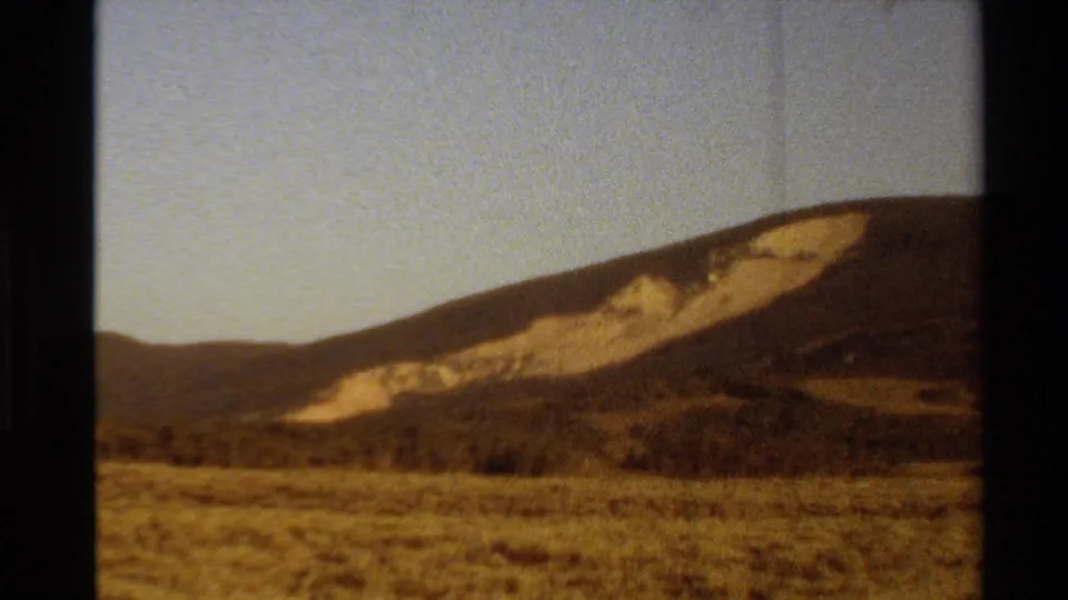 1980 Western U.S. home-movie frame of open grassland beneath a dark hill marked by pale exposed slope areas.