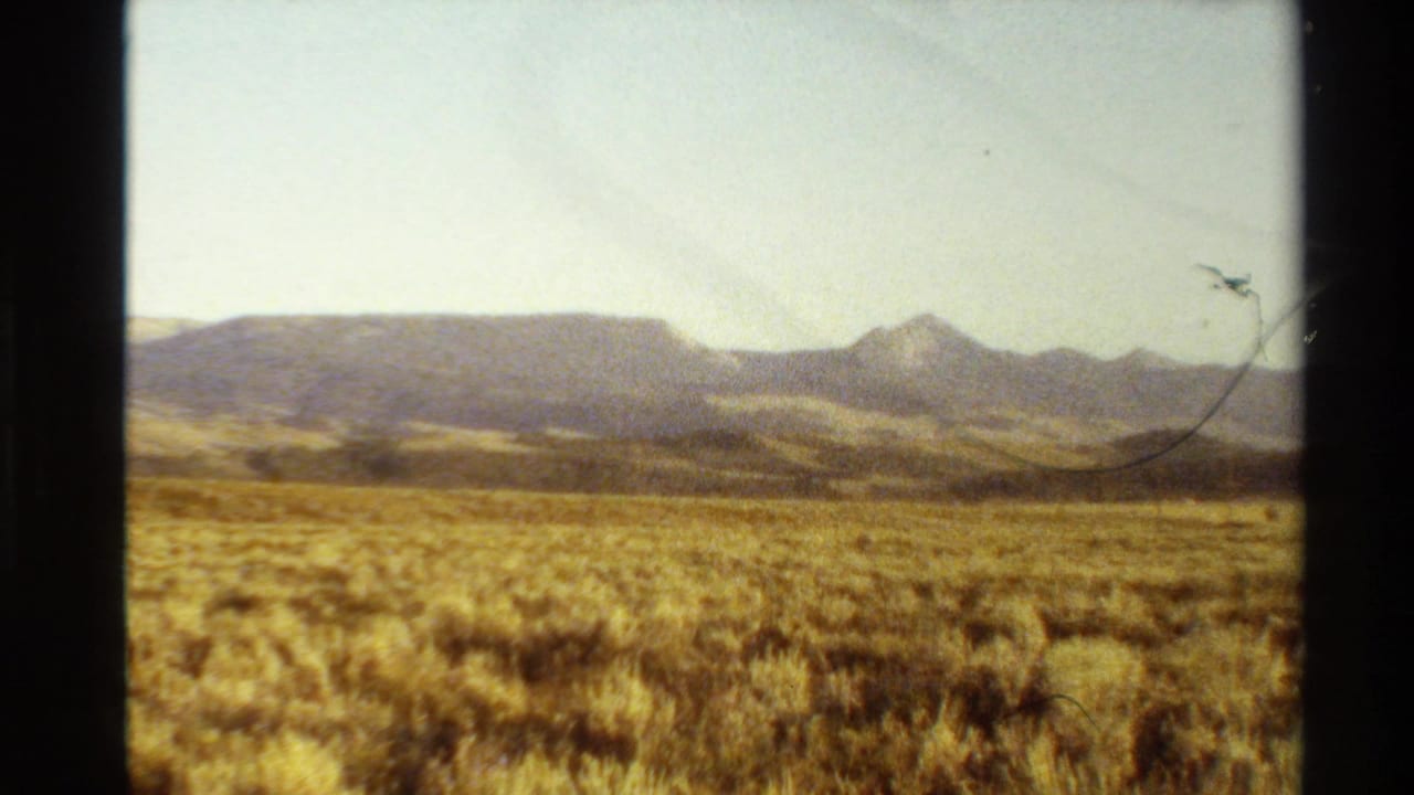 1980 Western USA 8mm frame showing dry grassland, distant ridges, pointed peaks, and a hazy pale sky within dark film borders.