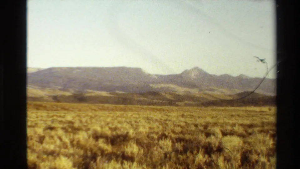 1980 Western USA 8mm frame showing dry grassland, distant ridges, pointed peaks, and a hazy pale sky within dark film borders.