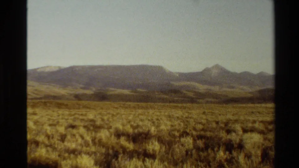 1980 Western USA landscape frame with golden grass in the foreground, low ridges, distant peaks, and soft haze across the sky.