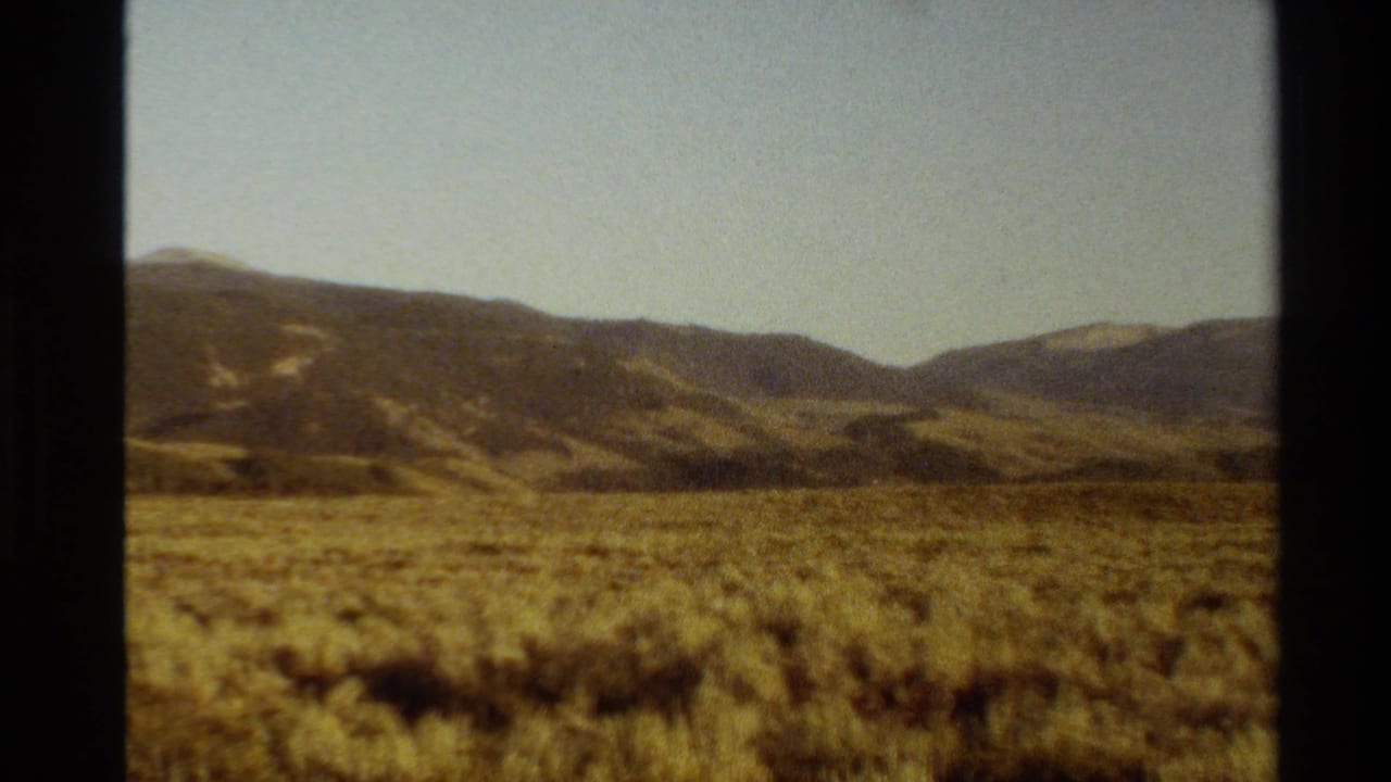 Western U.S. home-movie frame with sunlit meadow, rounded hills, and dark 8mm frame edges visible.