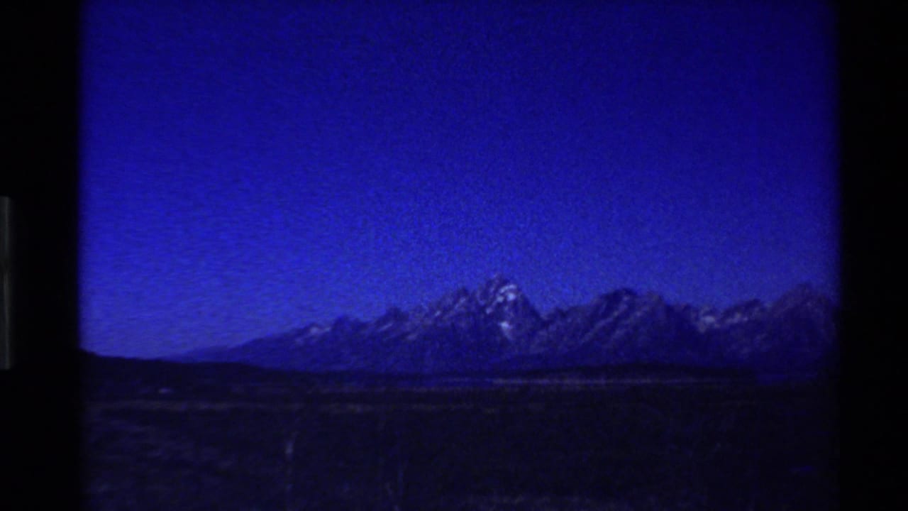 Underexposed 1980s 8mm frame of a Western U.S. mountain range beyond dark grassland, with a deep blue sky and heavy film borders.