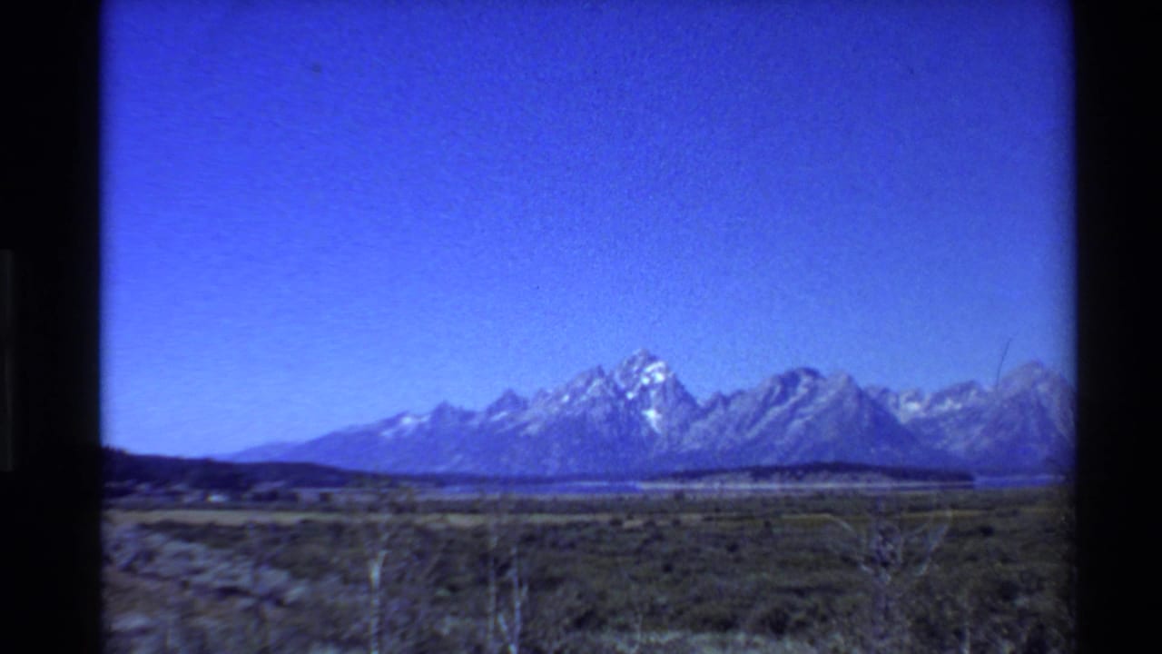 Brightened 1980s 8mm view of open Western U.S. grassland leading toward a distant snow-capped mountain range under clear sky.