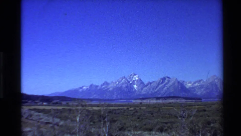 Brightened 1980s 8mm view of open Western U.S. grassland leading toward a distant snow-capped mountain range under clear sky.