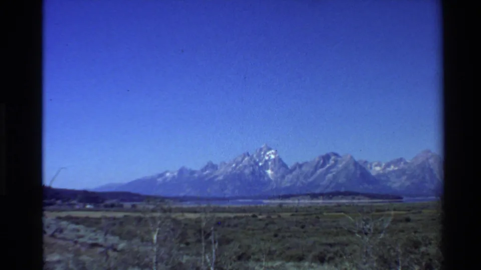 1980s 8mm landscape frame showing a wide grassy plain, a possible distant water or valley band, and snow-covered Western U.S. peaks.